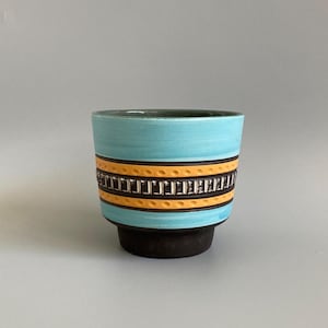 May include: A ceramic plant pot with a turquoise body, featuring an orange band and a dark brown base. The orange band has a repeating rectangular design. Suitable for indoor or outdoor use, it is a decorative item.