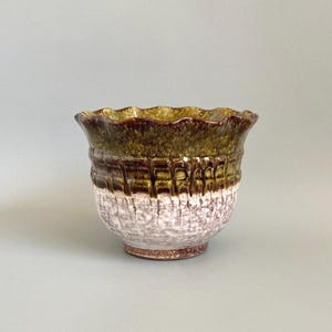 May include: A ceramic bowl featuring a scalloped rim and a two-tone glaze. The upper portion displays a deep olive green with vertical drip patterns, while the lower section transitions into a textured, mottled white and brown finish.