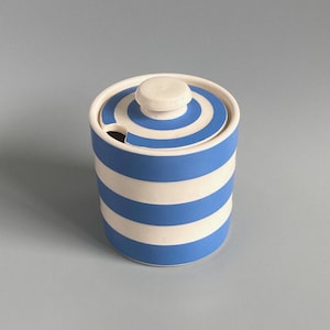 May include: A cylindrical ceramic jar featuring horizontal blue and white stripes. The matching lid has a central knob and a small cutout notch on the side, designed to accommodate a spoon. The container has a smooth, matte finish throughout.