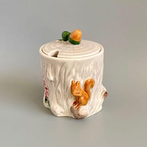 May include: A ceramic jar shaped like a tree stump with a textured white surface. A small orange squirrel is sculpted on the front. The lid features a moulded acorn handle and a notch for a spoon, with pink floral details visible on the side.