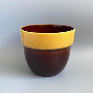 May include: A ceramic plant pot featuring a two-tone design with a glossy mustard yellow upper section and a deep, dark brown lower section. The vessel has a wide, open top with a smooth, rounded rim and a tapered base, showcasing a clean, simple form.