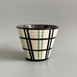 May include: A ceramic cup with a cream base, featuring a grid pattern of black and light green stripes. The rim and interior are dark brown. The cup has a slightly flared shape.