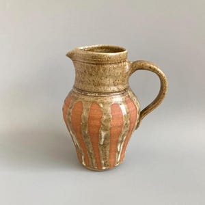 May include: A handmade ceramic jug with a handle and spout. The jug features vertical orange stripes against a beige background. The top and handle are a darker beige colour. A rustic, decorative piece.