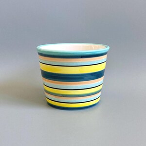 May include: A ceramic planter with a tapered cylindrical shape featuring horizontal stripes in shades of bright yellow, deep teal, light blue, and soft peach. The rim is finished in a solid turquoise glaze, and the interior is white.