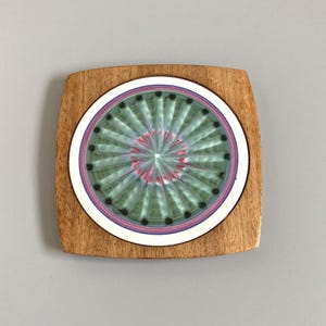 May include: A square wooden coaster with a circular ceramic insert. The insert has a radial design in green and pink, with black dots and a white and purple border. The coaster is made of light brown wood.