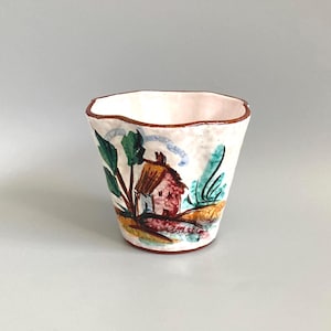 May include: A hand-painted ceramic cup with an irregular, wavy rim. The exterior features a colourful illustration of a small house with a brown roof, surrounded by green foliage and yellow ground accents, all set against a white background.