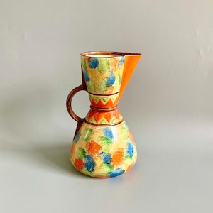 May include: A decorative ceramic jug with a distinctive shape and vibrant colours. The jug has a rounded base and a triangular spout. It is adorned with a pattern of blue, orange, and green spots. The top section is bright orange.