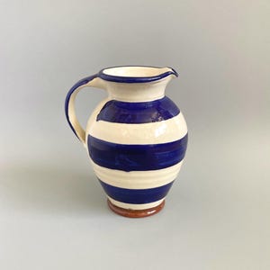 May include: A ceramic jug with a cream-coloured body and horizontal blue stripes. The jug has a curved handle and a spout for pouring. The base is reddish-brown. The jug is a decorative item.