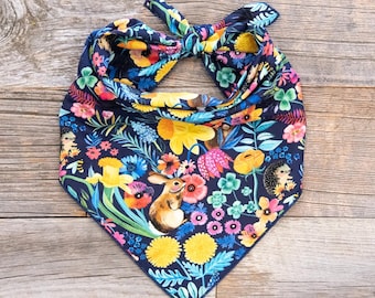 May include: A navy blue bandana with a floral print featuring a rabbit, hedgehog, and daffodils. The bandana is tied in a knot at the top.