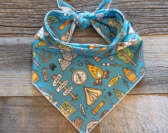 May include: A blue dog bandana with a camping theme. The bandana features illustrations of a compass, campfire, tent, trees, and other camping-related icons. The bandana is tied at the top with a white border.