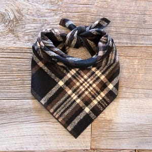 May include: A plaid dog bandana in shades of black, brown, and cream. The fabric is knotted at the top, forming a triangle shape. The pattern features a classic plaid design, perfect for pet accessories.