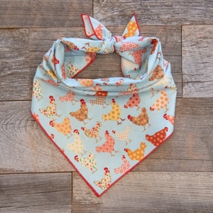 May include: A light blue dog bandana with a repeating pattern of colorful chickens. The bandana is tied at the top and has a red trim. The chickens are in various colors and patterns, creating a cheerful and whimsical design.