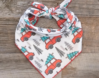 May include: White bandana with a red border and a repeating pattern of red cars carrying Christmas trees. The cars are driving through a forest of gray and green trees.