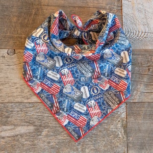 May include: A triangular pet bandana featuring a blue camouflage pattern with printed dog tags, American flags, and the text Honor, Respect, Commitment, Courage, Praise Our Heroes, and USAF. The fabric has a thin red border along the bottom edge.