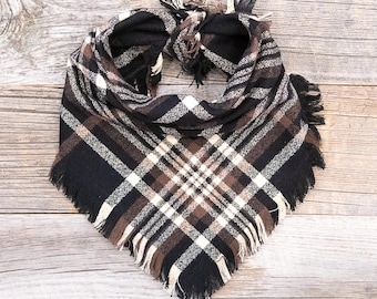 May include: A plaid scarf in a triangular shape, featuring a pattern of black, brown, and cream-colored squares. The scarf has a fringed edge and is displayed on a wooden surface.