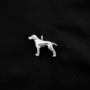 Könnte beinhalten: Ein polierter, silberfarbener Metallanhänger in Form eines stehenden Hundes im Profil. Die Hunde-Figur hat eine glatte, reflektierende Oberfläche und einen kleinen, runden Verbindungsring auf dem Rücken, um sie an einer Kette zu befestigen.