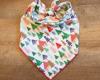 May include: White bandana with a repeating pattern of colorful Christmas trees in red, green, gold, and blue. The bandana has a red border and is tied in a knot at the top. The fabric is cotton.