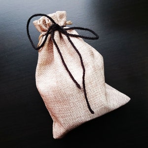 Peut inclure: Un petit sac en toile de jute beige avec des cordons de serrage noirs nou&eacute;s en un n&oelig;ud. Le sac est pos&eacute; sur une surface noire.