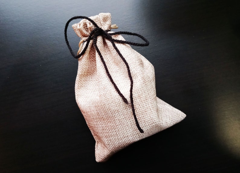 Peut inclure: Un petit sac en toile de jute beige avec des cordons de serrage noirs nou&eacute;s en un n&oelig;ud. Le sac est pos&eacute; sur une surface noire.