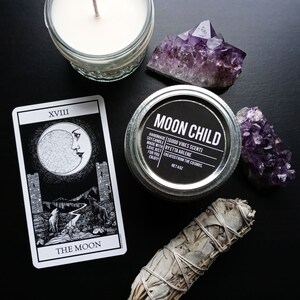 Peut inclure: Une carte de tarot noir et blanc repr&eacute;sentant La Lune (XVIII) avec un croissant de lune, deux tours et un loup. Un b&acirc;tonnet de sauge blanche, un cristal d'am&eacute;thyste violet, une bougie dans un bocal en verre et un bocal de bougie &eacute;tiquet&eacute; "Moon Child" sont &eacute;galement pr&eacute;sents sur l'image.