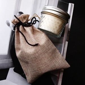 Peut inclure: Un petit pot en verre transparent avec un couvercle argent&eacute; rempli d'une bougie blanche. Le pot est pos&eacute; sur un sac en toile de jute brun avec des liens en ficelle noire.