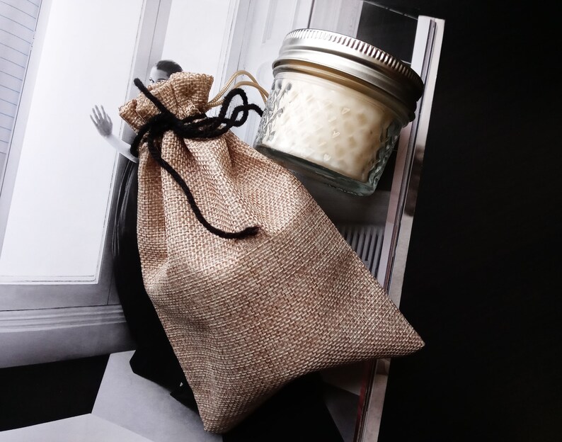 Peut inclure: Un petit pot en verre transparent avec un couvercle argent&eacute; rempli d'une bougie blanche. Le pot est pos&eacute; sur un sac en toile de jute brun avec des liens en ficelle noire.