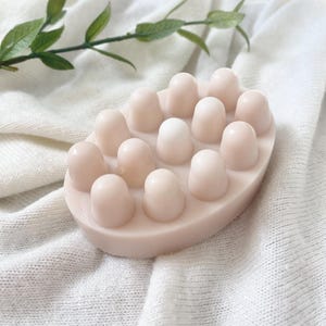 May include: A light pink, oval-shaped massage soap with rounded, raised nodules. The soap is set against a white, textured fabric, with a sprig of green leaves in the background. The soap's design is intended for massage.