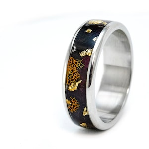 Puede incluir: Un anillo de banda de plata con una incrustación negra y dorada. La incrustación presenta un patrón de escamas doradas y una resina oscura, casi púrpura.
