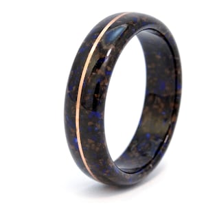 May include: A dark ring with a speckled pattern of brown and blue, and a thin copper-colored stripe down the center. The ring is set against a white background, highlighting its unique design.