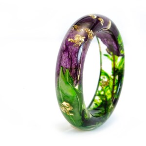 K&ouml;nnte beinhalten: Ein klarer Armreif aus Kunstharz mit lila Bl&uuml;ten, gr&uuml;nen Bl&auml;ttern und Goldblatt-Akzenten. Das botanische Design ist in dem transparenten Harz eingeschlossen und schafft ein einzigartiges Schmuckst&uuml;ck. Das Armband hat eine kreisf&ouml;rmige Form.