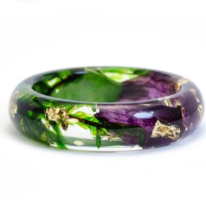K&ouml;nnte beinhalten: Ein klarer Harzring mit eingebetteten gr&uuml;nen Bl&auml;ttern, lila Bl&uuml;tenbl&auml;ttern und Goldblattflocken. Der Ring ist ein dickes Band, das die eingeschlossenen Naturelemente zeigt. Das Design ist vor einem wei&szlig;en Hintergrund.