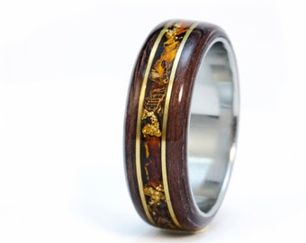 Puede incluir: Un anillo de madera marrón con una incrustación dorada y una banda plateada. El anillo tiene un diseño único con escamas de oro incrustadas en la madera.