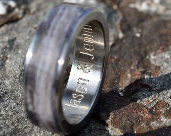 Puede incluir: Un anillo plateado con un patrón de vetas de madera y la inscripción "Jason & Jenna" grabada en el interior. El anillo está sobre un fondo de piedra gris texturizada.