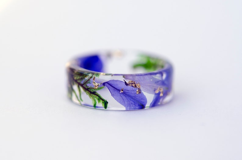 May include: A clear resin ring with embedded blue and purple flower petals, green foliage, and gold flakes. The ring has a wide band and is set against a white background. A unique piece of jewelry.