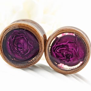 May include: A pair of wooden ear plugs featuring a dark purple rose design set within clear resin. The plugs have a natural wood grain finish. The rose is the central element, set against a white backdrop.