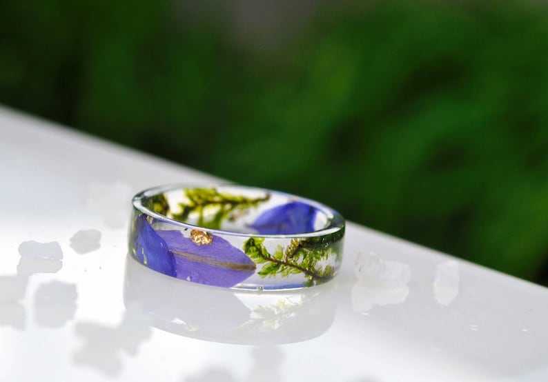 May include: A clear resin ring with a botanical design. The ring features blue petals, green moss, and gold flakes encased within the transparent band. The ring is set against a white surface with a blurred green background.