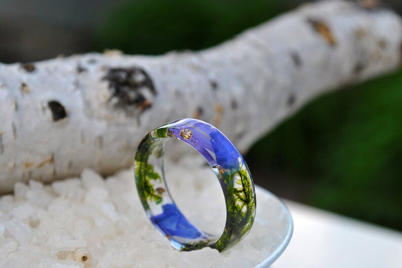 May include: A clear resin ring with a vibrant blue floral design and gold flakes. The ring features green foliage and is set against a backdrop of white crystals and a blurred natural background. A unique piece of jewelry.