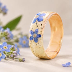 May include: A circular ring made of a light-colored, speckled material featuring embedded blue forget-me-not flowers. The band has a smooth, polished finish with a visible wood-like grain texture on the inner surface and a rounded outer profile.
