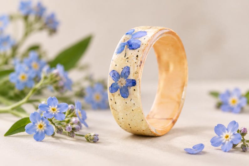 May include: A circular ring made of a light-colored, speckled material featuring embedded blue forget-me-not flowers. The band has a smooth, polished finish with a visible wood-like grain texture on the inner surface and a rounded outer profile.
