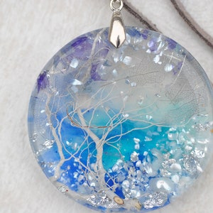 May include: A round, blue and white resin pendant with a silver bail. The pendant features a white tree branch design and iridescent glitter.