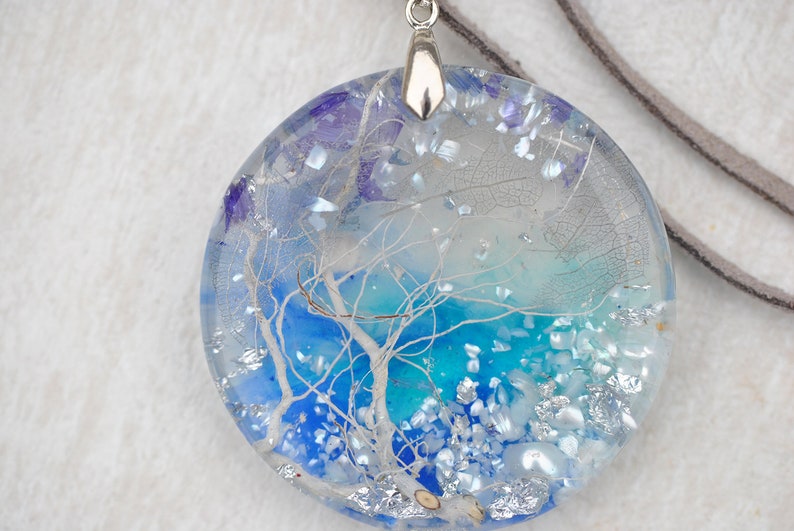 May include: A round, blue and white resin pendant with a silver bail. The pendant features a white tree branch design and iridescent glitter.