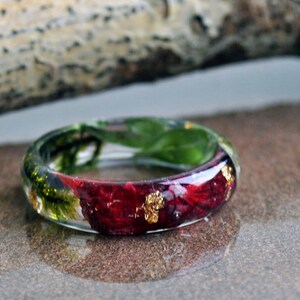 May include: A clear resin ring with embedded red flowers, green leaves, and gold flakes. The ring has a rounded shape and is set against a neutral background. A unique piece of jewelry.