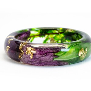 K&ouml;nnte beinhalten: Ein klarer Harzring mit einem botanischen Design. Der Ring hat lila Blumen, gr&uuml;ne Bl&auml;tter und Blattgoldflocken. Der Ring ist etwa 2,5 cm breit.