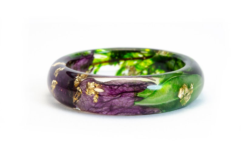 K&ouml;nnte beinhalten: Ein klarer Harzring mit einem botanischen Design. Der Ring hat lila Blumen, gr&uuml;ne Bl&auml;tter und Blattgoldflocken. Der Ring ist etwa 2,5 cm breit.