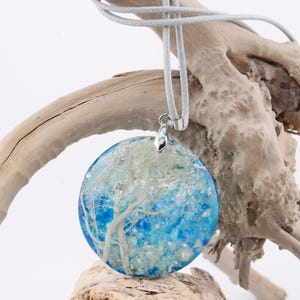 May include: A round blue and white resin pendant with a silver bail. The pendant features a white tree-like design inside the resin. The pendant is hanging from a gray cord on a piece of driftwood.