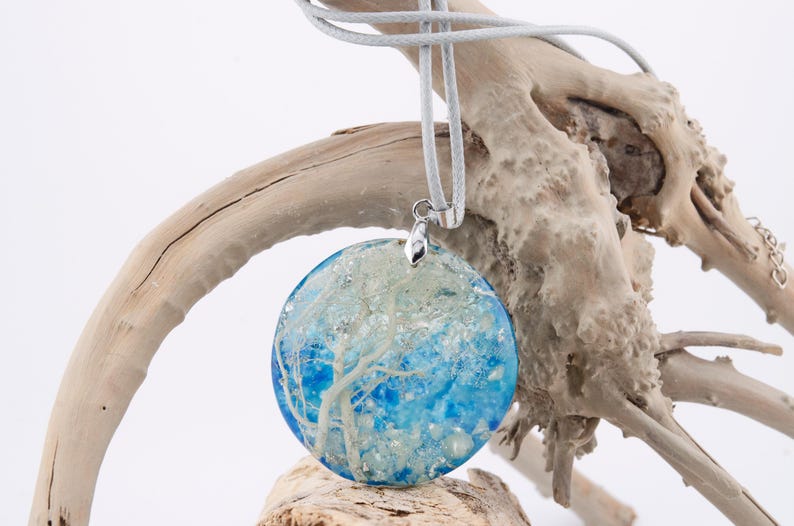 May include: A round blue and white resin pendant with a silver bail. The pendant features a white tree-like design inside the resin. The pendant is hanging from a gray cord on a piece of driftwood.