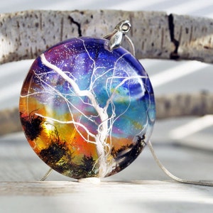 May include: A round pendant necklace with a white tree silhouette inside a clear resin dome. The tree is surrounded by a colorful nebula-like background with shades of blue, purple, orange, and yellow.