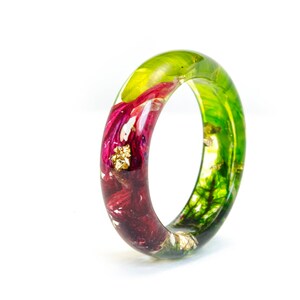 May include: A clear resin ring with embedded flowers and gold flakes. The ring features a vibrant mix of colors, including deep red, green, and gold. The ring is a unique piece of jewelry.