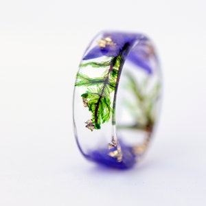 May include: A clear resin ring with a band of purple and embedded with green fern fronds, purple flowers, and gold flakes. The ring is a unique piece of jewelry.