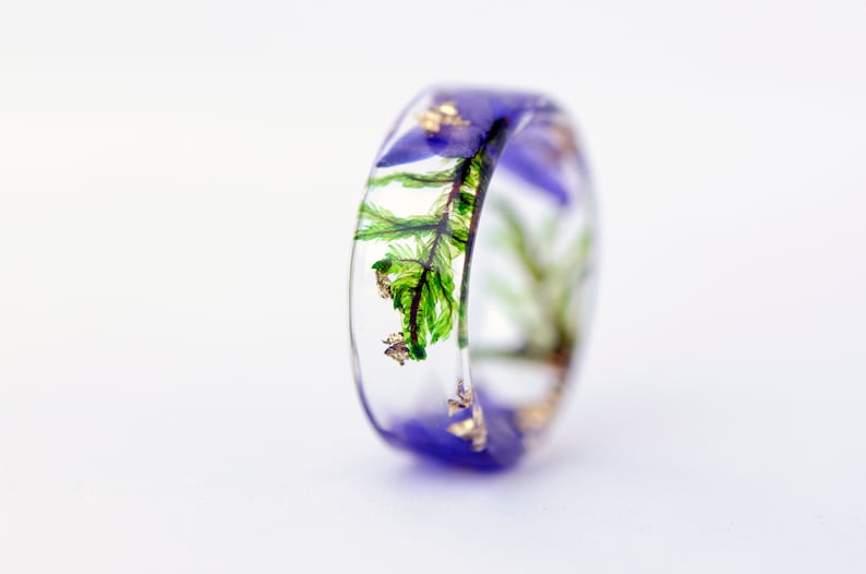 May include: A clear resin ring with a band of purple and embedded with green fern fronds, purple flowers, and gold flakes. The ring is a unique piece of jewelry.